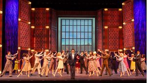 A group musical number with a man in a suit in the middle