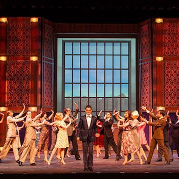 A group musical number with a man in a suit in the middle