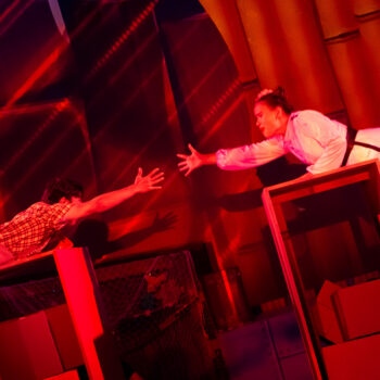 Two actors reaching across red-lit stage
