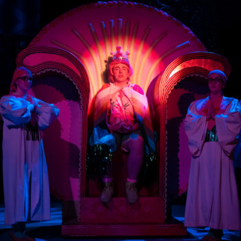 Performer crowned on ornate throne, robed attendants