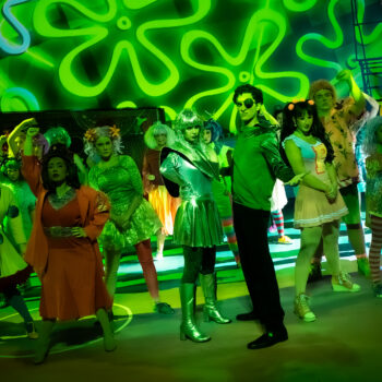 Colorful costumed cast on green-lit stage