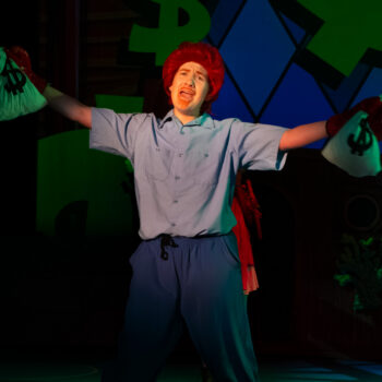 Performer with red wig holding dollar bags